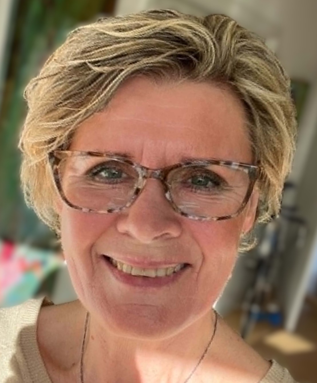 Smilende kvinde med kort blondt hår, perleøreringe og halskæde, fotograferet mod lys baggrund.